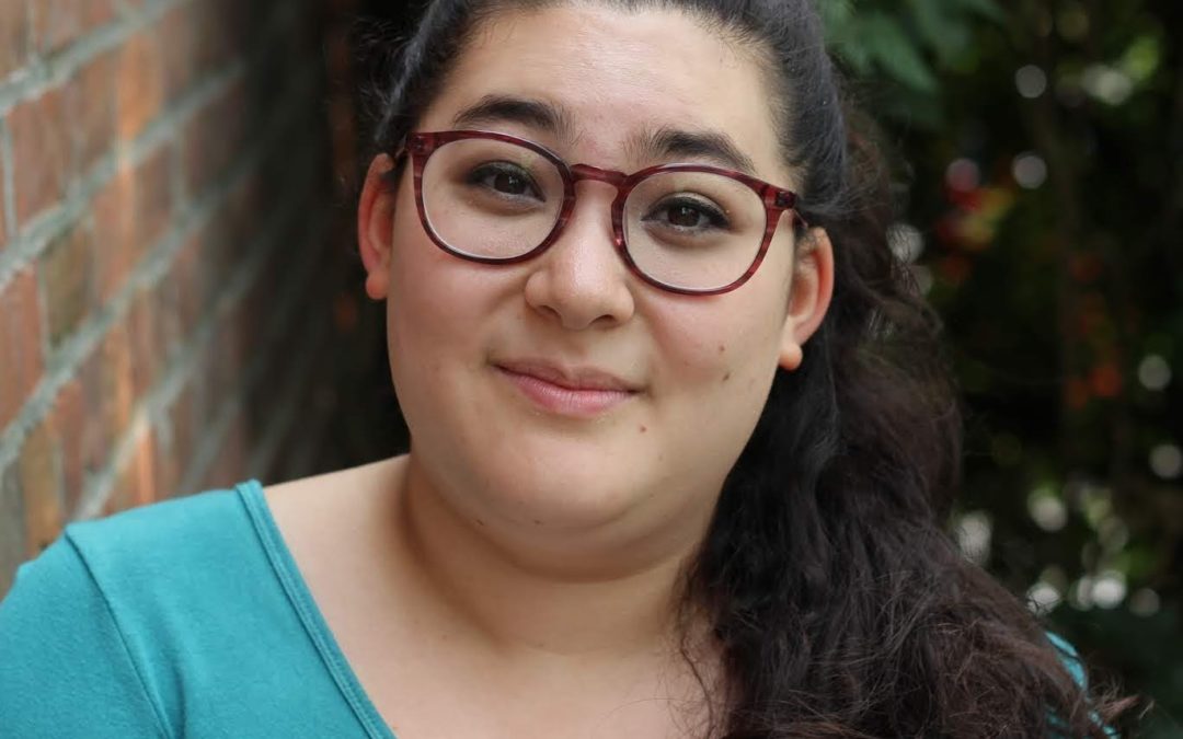 In Hannah's headshot, she stands in front of a brick wall, wearing a longsleeved aqua shirt. Her long dark hair is in a pony tail and she is wearing glasses. She smiles at the camera.
