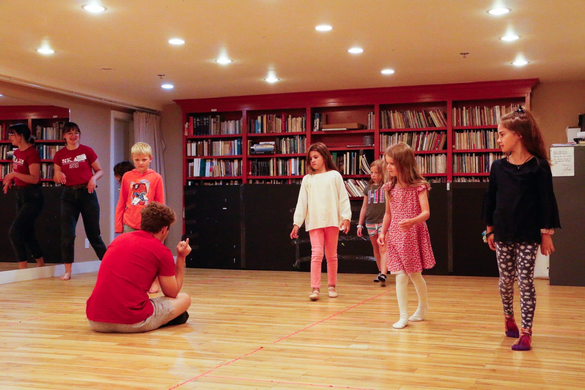 A group of kids sneak past their teacher in a studio room, as part of a game.